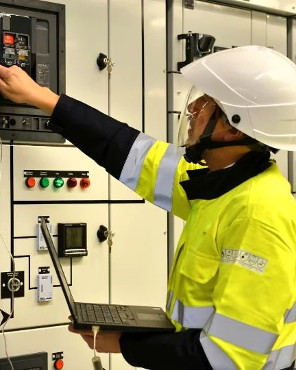 Técnico con casco, careta y ropa de alta visibilidad operando el interruptor de un tablero de baja tensión.